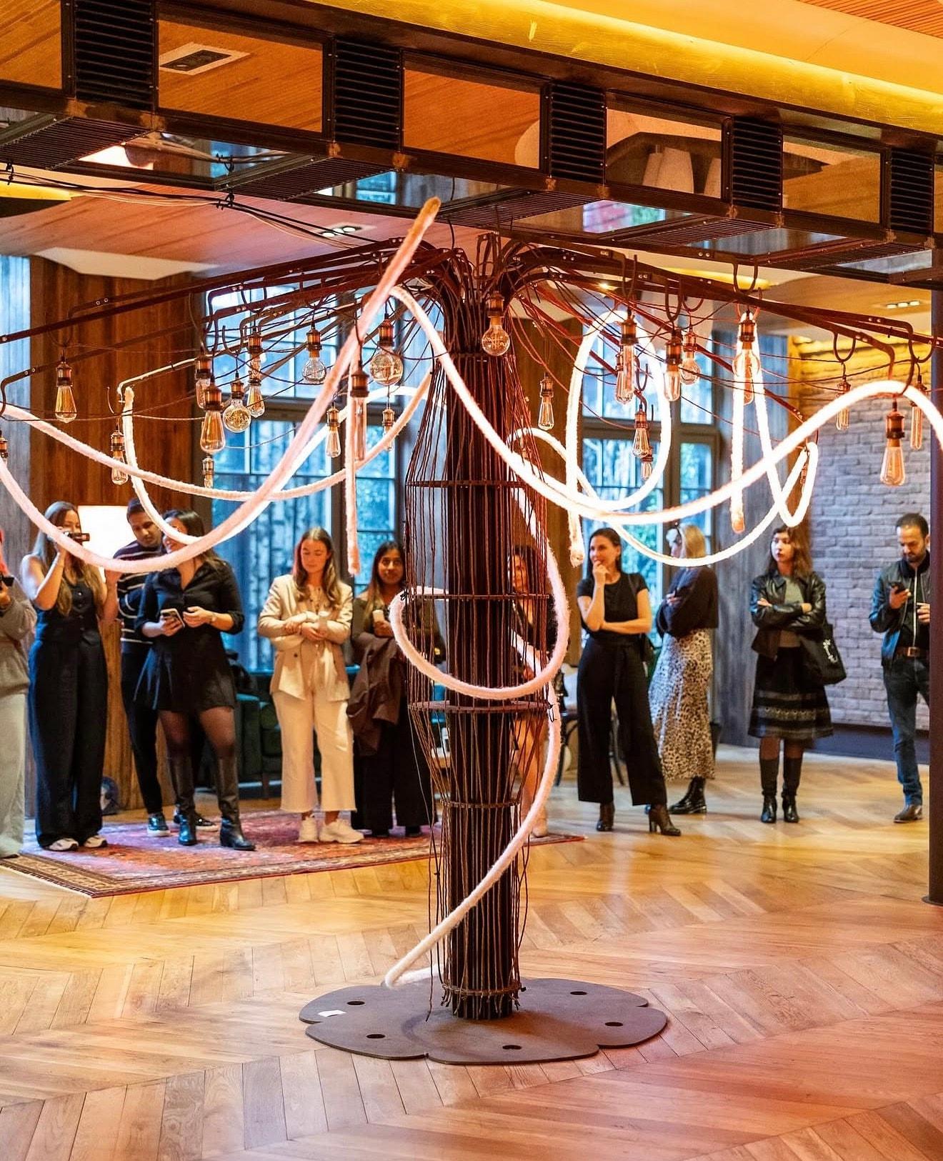 Glowing tree-like light installation with visitors gathered around it at the Symbiosis exhibition, Cicek Gallery London